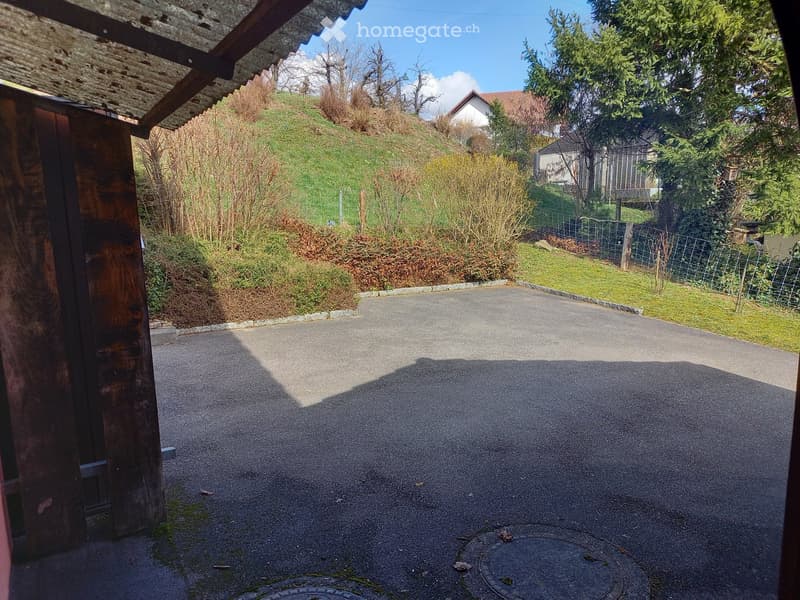 Driveway leading up to a house with a grassy slope and trees in the background.