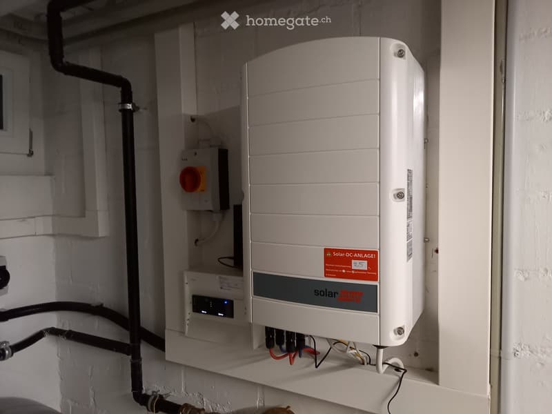 A solar inverter and circuit breaker mounted on a wall in a utility space, with visible wiring and connections.