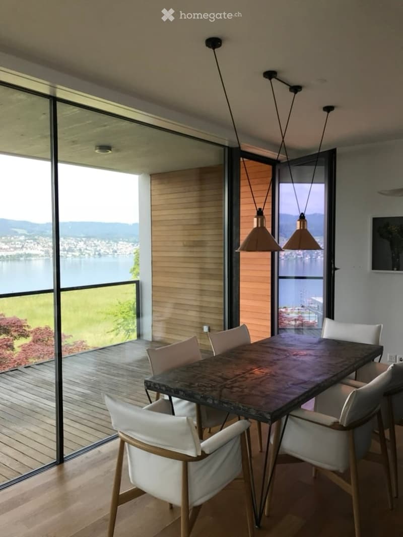 Modern dining area with a dark wooden table, white chairs, and large windows offering a scenic view of a lake and mountains.