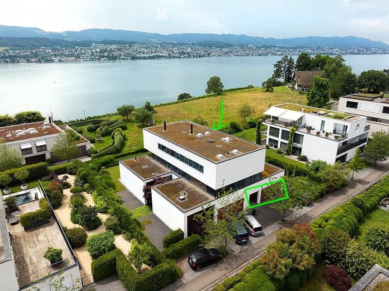 Aerial view of a modern residential complex with multiple buildings, surrounded by landscaped gardens, parking areas, and a scenic lake view in the background.