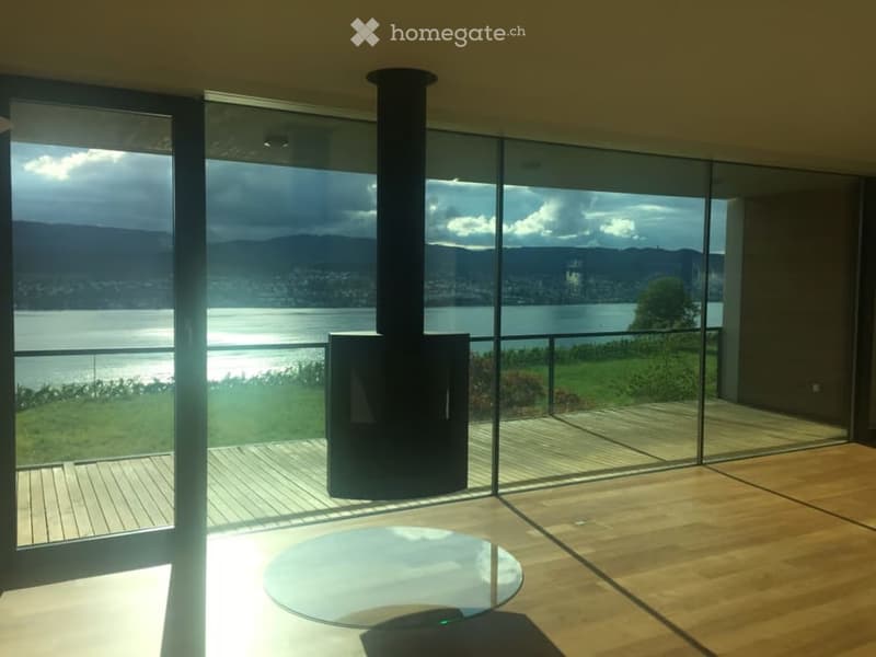 Modern living room with large glass walls offering a scenic view of a lake and mountains, featuring a round glass table and a central fireplace.