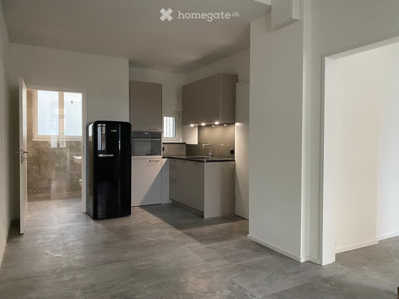 Modern kitchen with grey cabinets, a black refrigerator, a sink with a faucet, and an oven. The space features tiled flooring and ample natural light from a window.