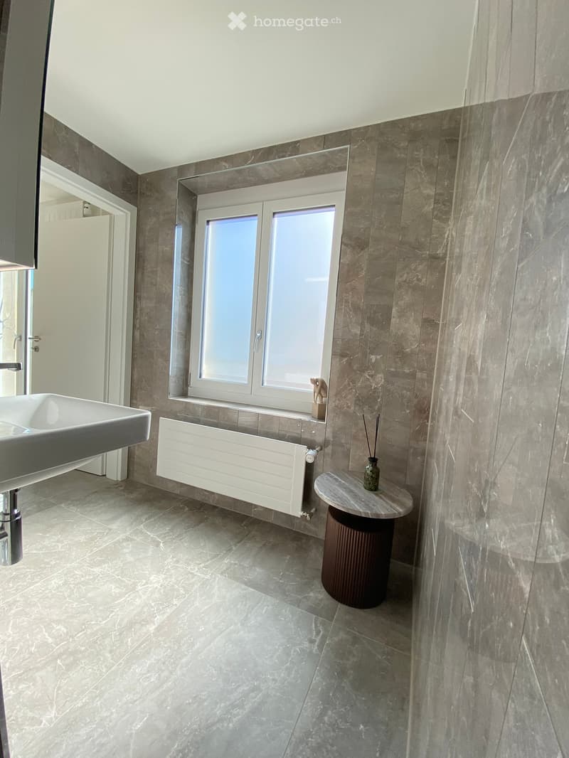 Modern bathroom with marble tiles, a white sink, a small round stool, a vase with sticks, and a radiator under a large window.