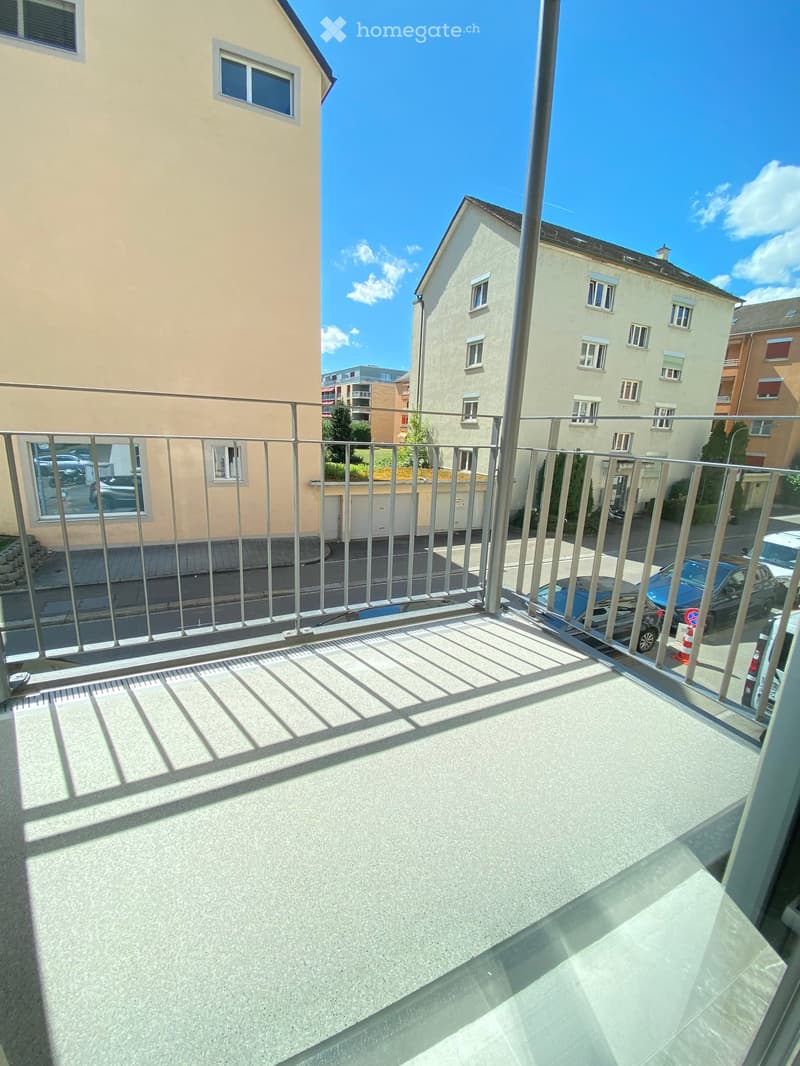 Balcony with metal railing, overlooking a residential area with buildings and parked cars.