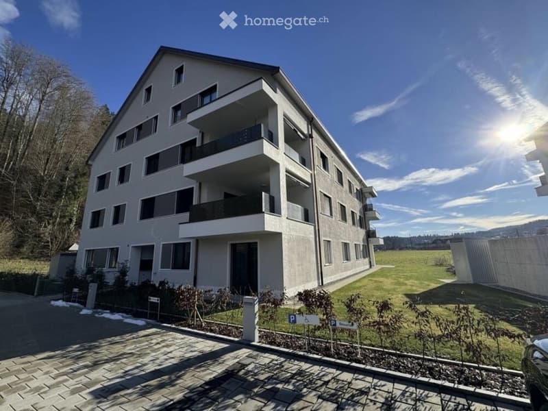 Modern multi-story residential building with balconies, parking area, and landscaped surroundings.