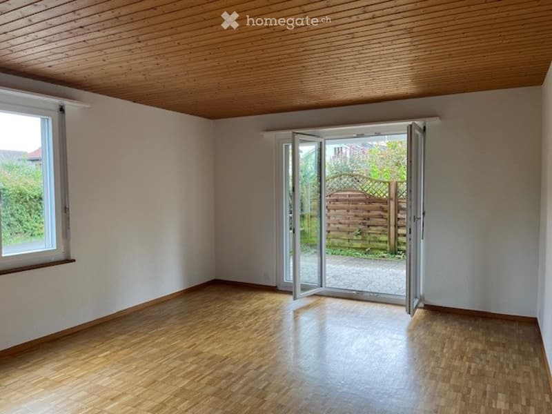 Empty room with wooden flooring, white walls, large window, and sliding glass door leading to a garden area.