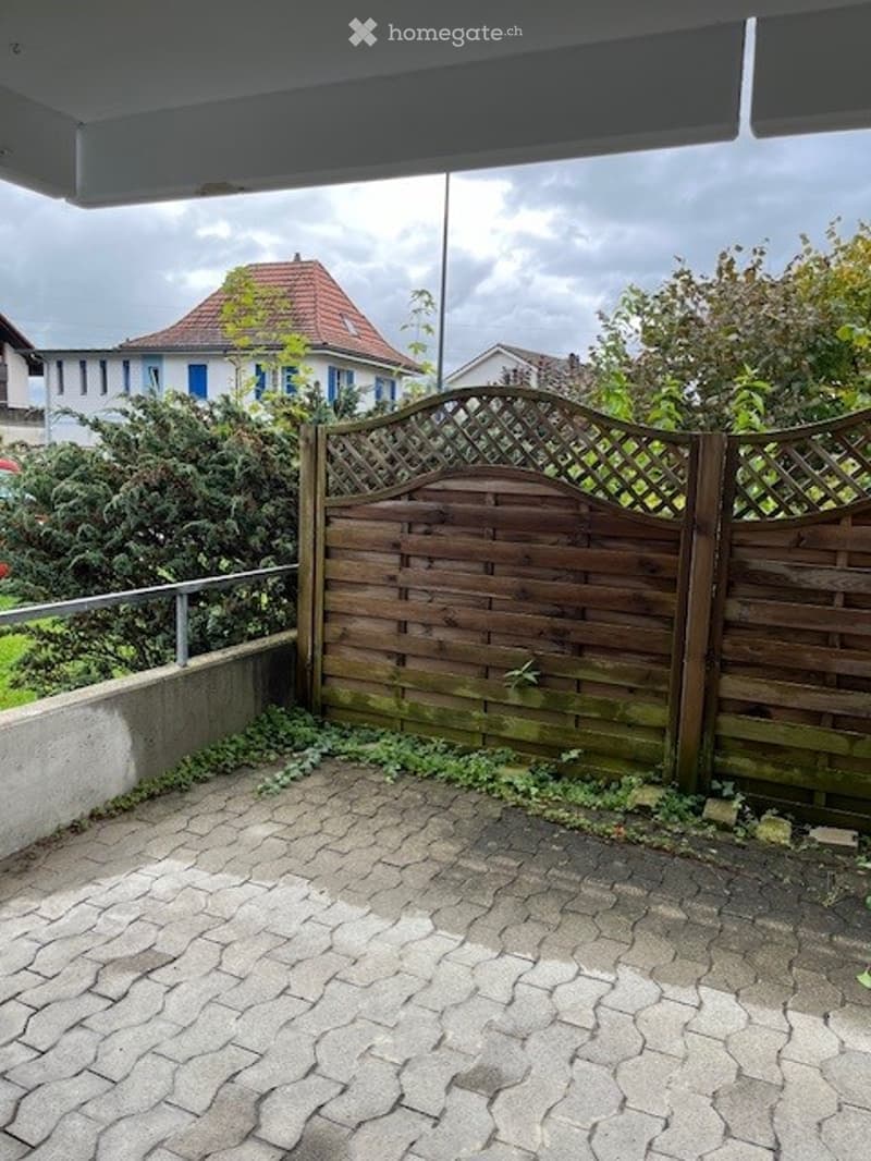 A paved outdoor area enclosed by a wooden lattice fence, with a house visible in the background.