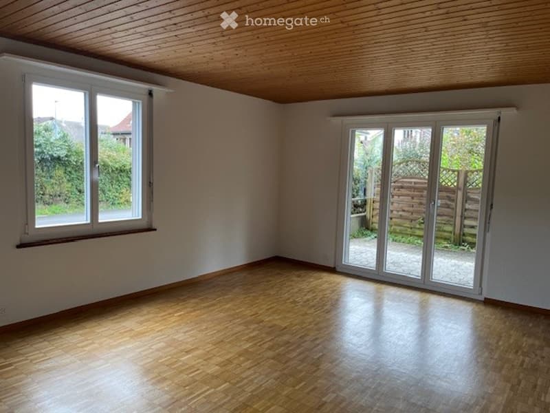 Empty room with wooden flooring, white walls, large windows, and a sliding glass door leading to an outdoor area.