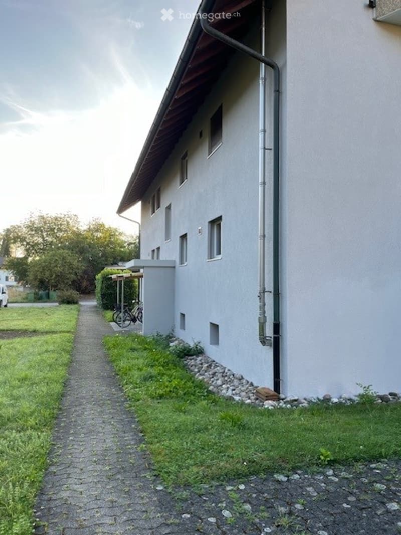 A modern, two-story building with a flat roof and white exterior. The building has multiple windows and a small covered entrance area. A paved pathway leads to the entrance, flanked by grass and small shrubs. A bicycle is parked near the entrance, and the