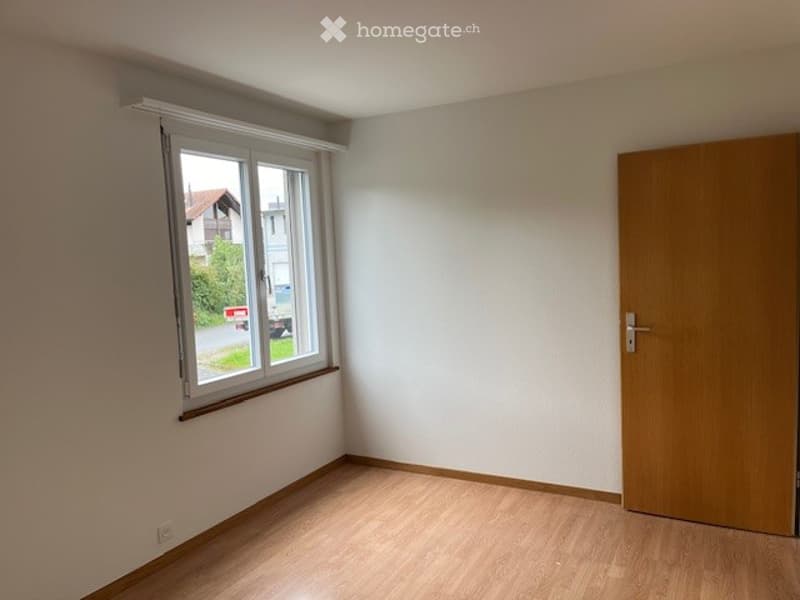 Empty room with light wooden flooring, white walls, a large window with white frames, and a wooden door.