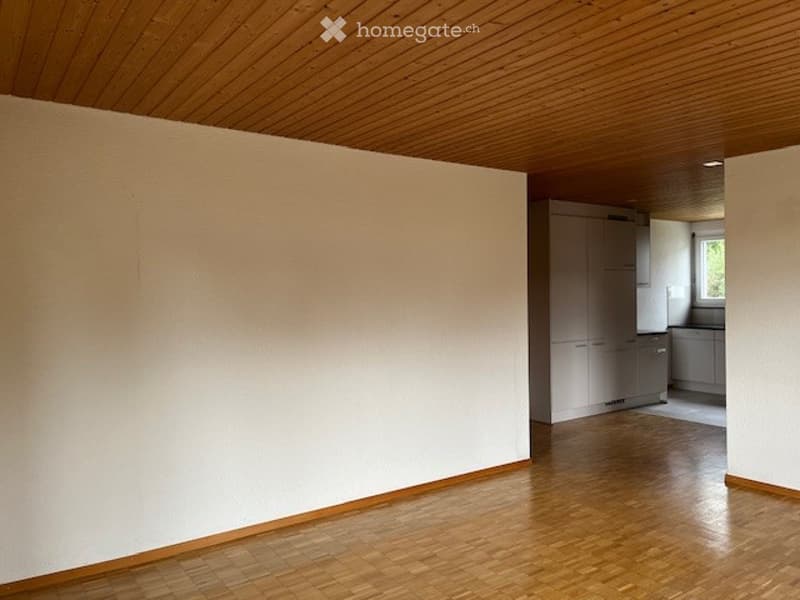 Empty room with wooden flooring and white walls, connected to a small kitchen area with cabinets and a window.