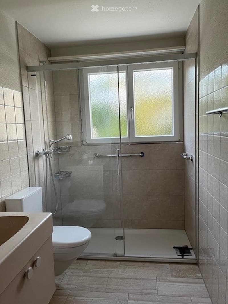 Modern bathroom with a glass-enclosed shower, toilet, and sink. The shower has a fixed glass door and a frosted window above it. The walls are tiled in beige, and the floor is made of light-colored tiles.