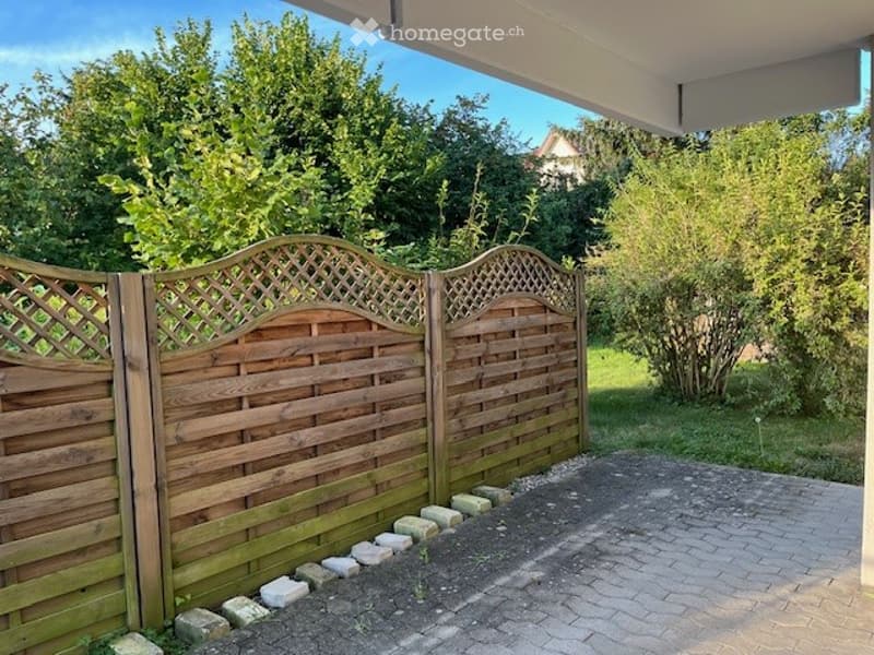 Wooden fence with decorative lattice and arched top, stone border, paved driveway, lush greenery, and trees in the background.