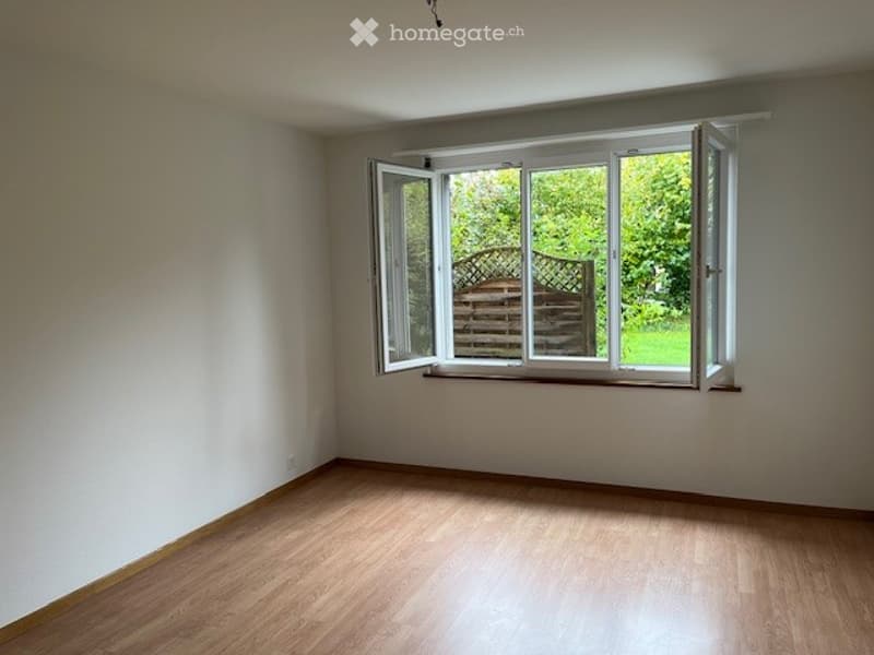 Empty room with wooden flooring, white walls, and large open window showing greenery outside.