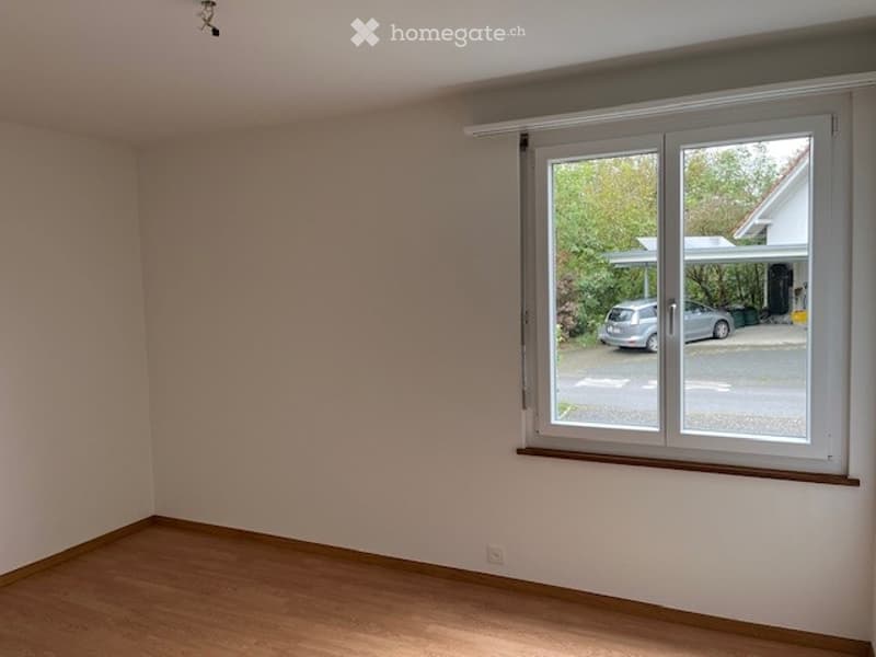 Empty room with wooden flooring, white walls, and a large window with blinds, showing a view of a driveway and parked car.