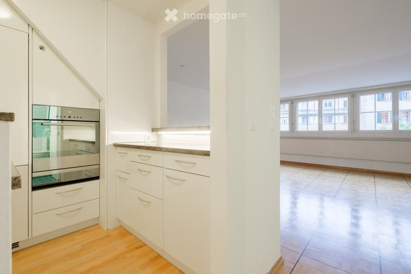 Modern kitchen with white cabinets, stainless steel oven, and granite countertop. Adjacent unfurnished room with large windows and tiled floor.
