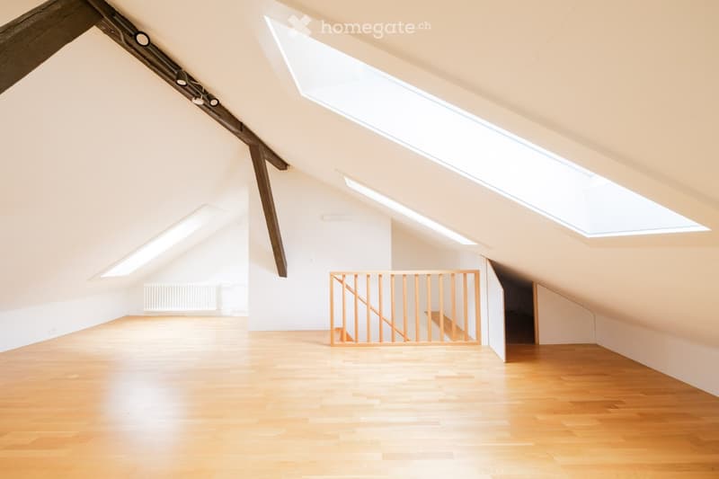 Bright, empty attic space with wooden flooring, white walls, exposed wooden beams, and skylights allowing natural light to fill the room. A simple wooden staircase leads to another area.
