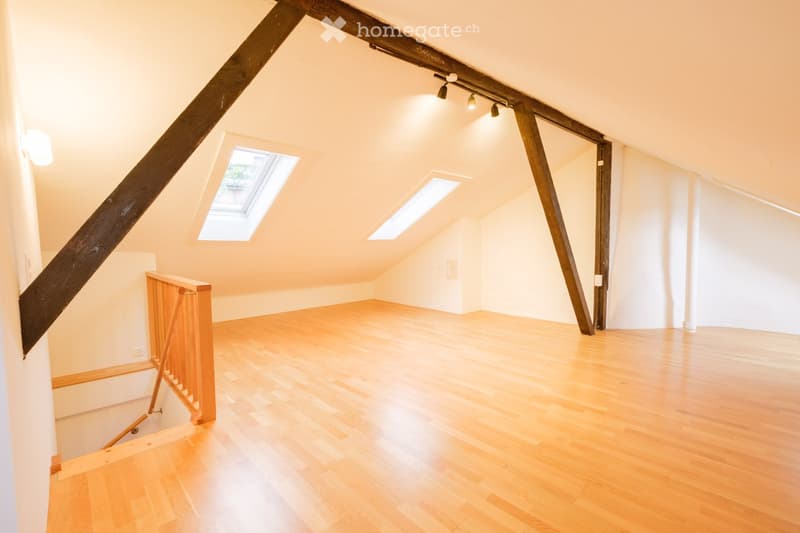 Empty attic room with wooden floors, exposed wooden beams, skylights, and a small staircase leading down.