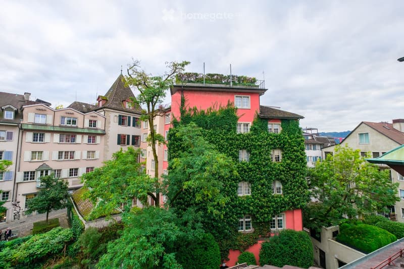 A view of a multi-story residential building with a pink facade, multiple windows, and greenery on the roof and surrounding area.