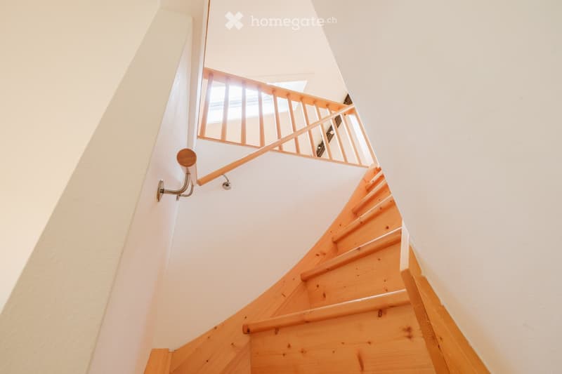 Spiral staircase with wooden steps and a wooden handrail, white walls, and a small window at the top.