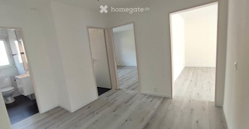 Empty room with light wooden flooring, white walls, and several open doors leading to adjacent spaces. No furniture or equipment is present.