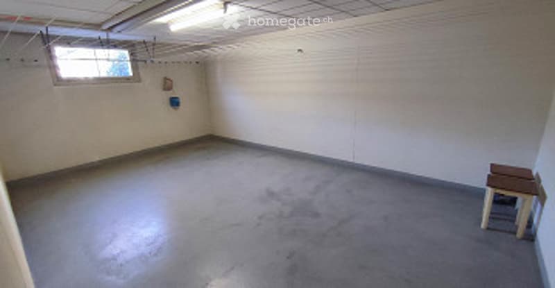 Empty room with concrete floor, white walls, single window, and a small wooden stool in the corner.