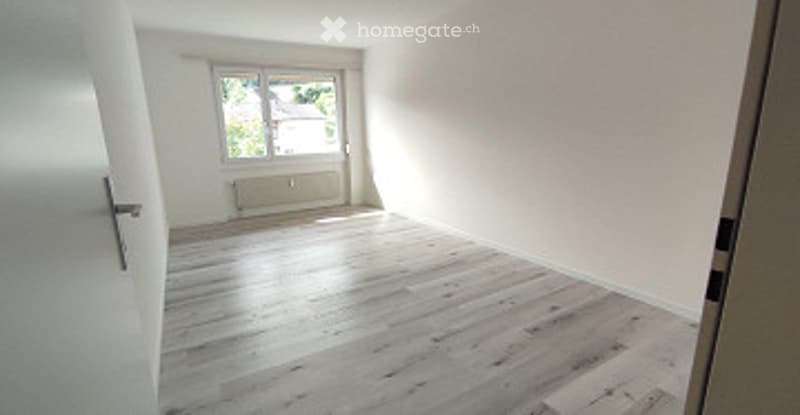 Empty room with light wooden flooring, white walls, and a window with a view of another building.