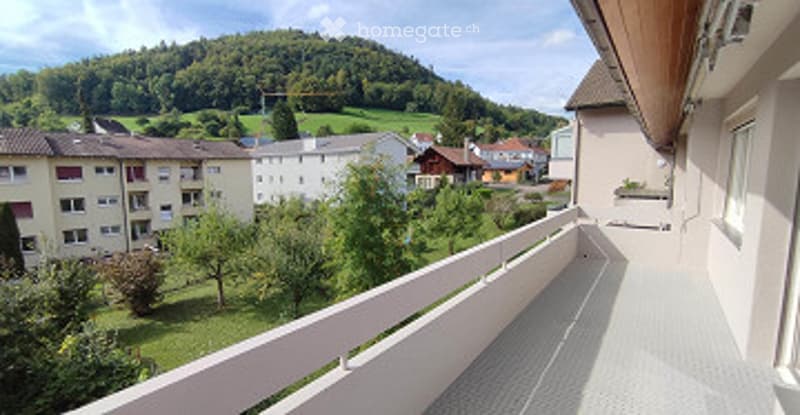 Balcony with white railing, tiled floor, overlooking a green landscape with houses and trees.