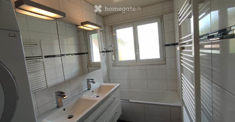 Modern bathroom with white tiled walls and floor, double sink with chrome faucets, bathtub, and large windows providing natural light.