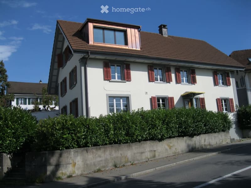 Two-story house with white walls, red shutters, brown tiled roof, and a small dormer window. A low hedge borders the front yard.