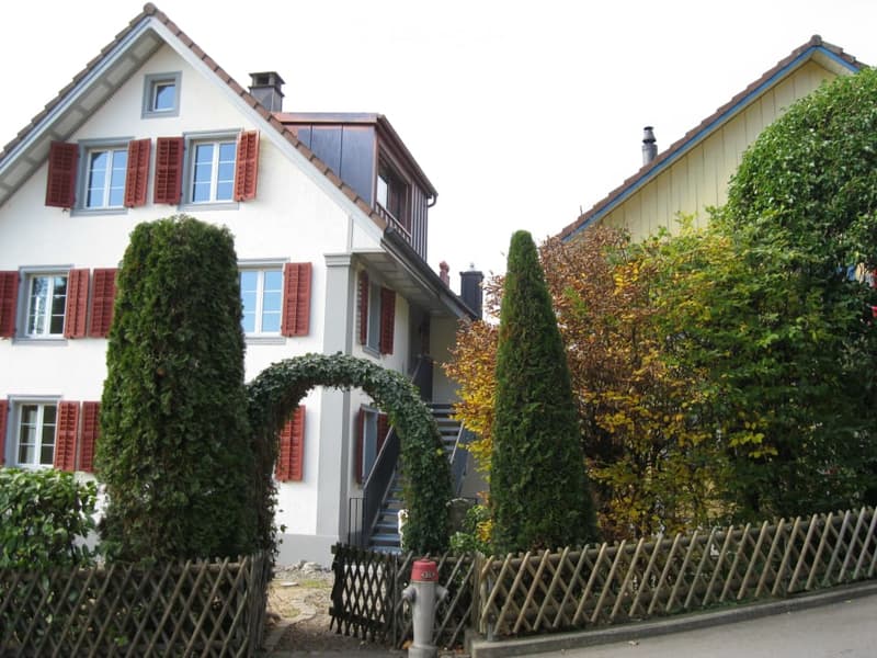 Two-story white house with red shutters, a small staircase leading to the entrance, and a wooden fence with greenery.