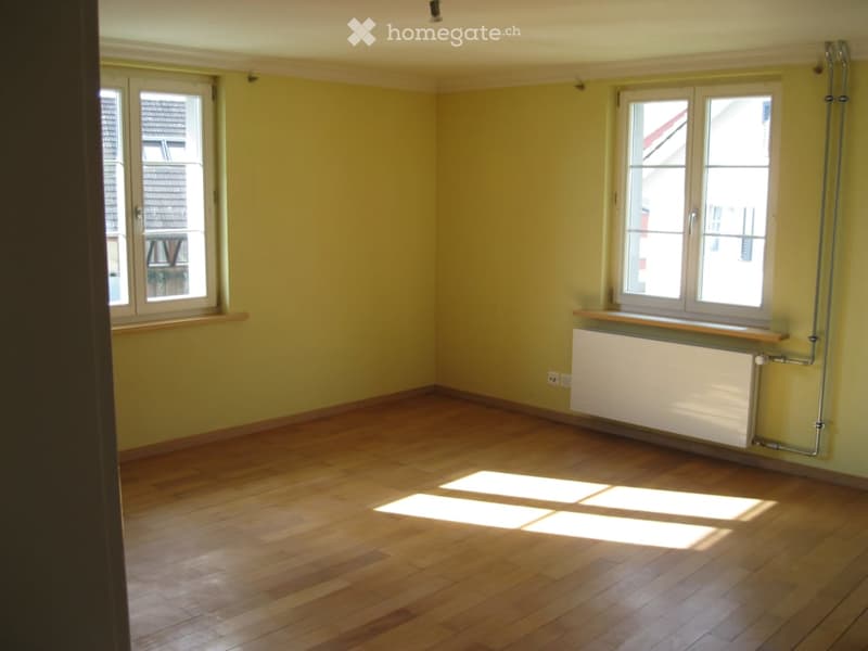 Empty room with light yellow walls and wooden flooring, featuring two windows with white frames and a radiator below one window.