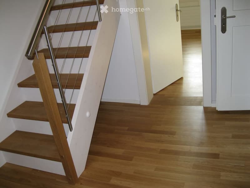 A modern hallway with wooden flooring and a staircase featuring wooden steps and a metal railing. The hallway has white walls and a closed white door at the end.