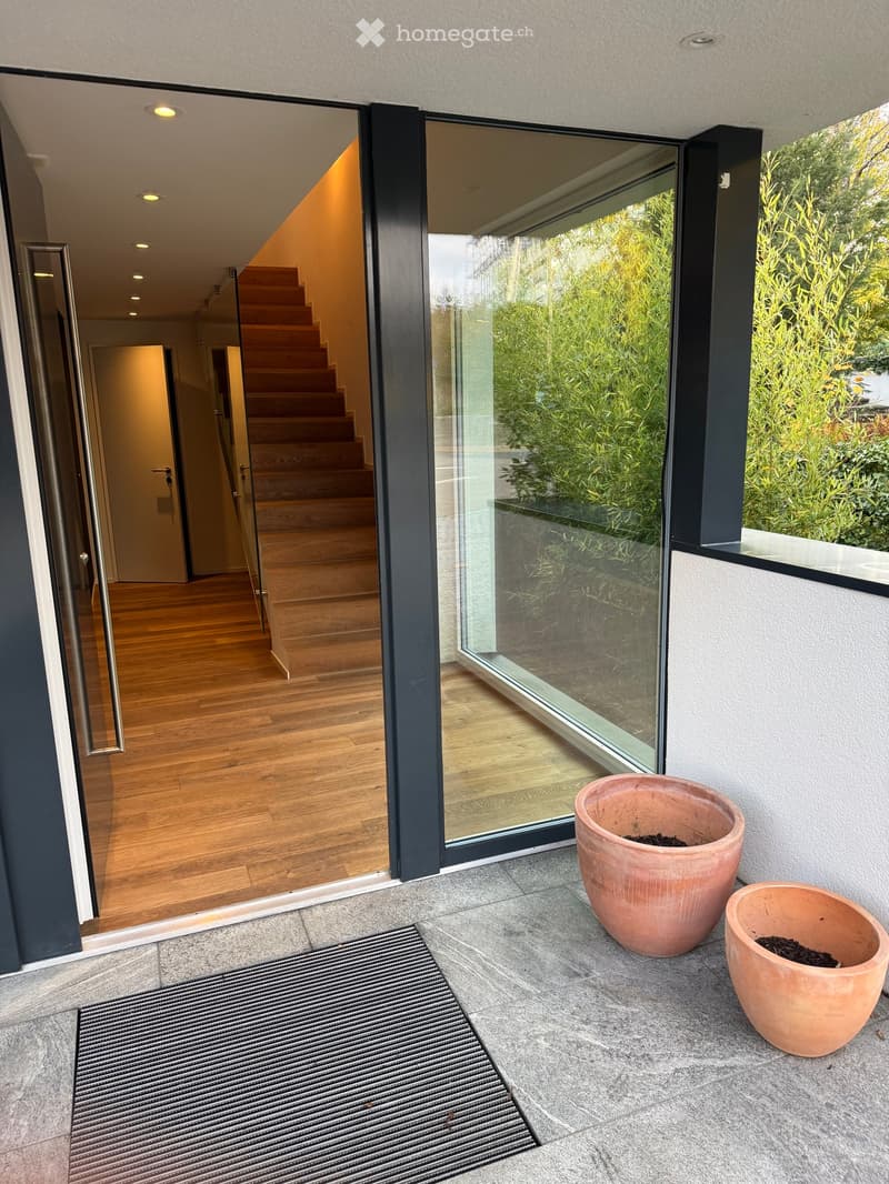 Modern outdoor area with large glass sliding doors leading to a wooden interior, two large terracotta pots on a stone patio, and a metal doormat.