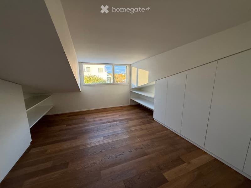 Empty attic room with wooden flooring, white built-in shelving, and large windows offering a view of the outside.