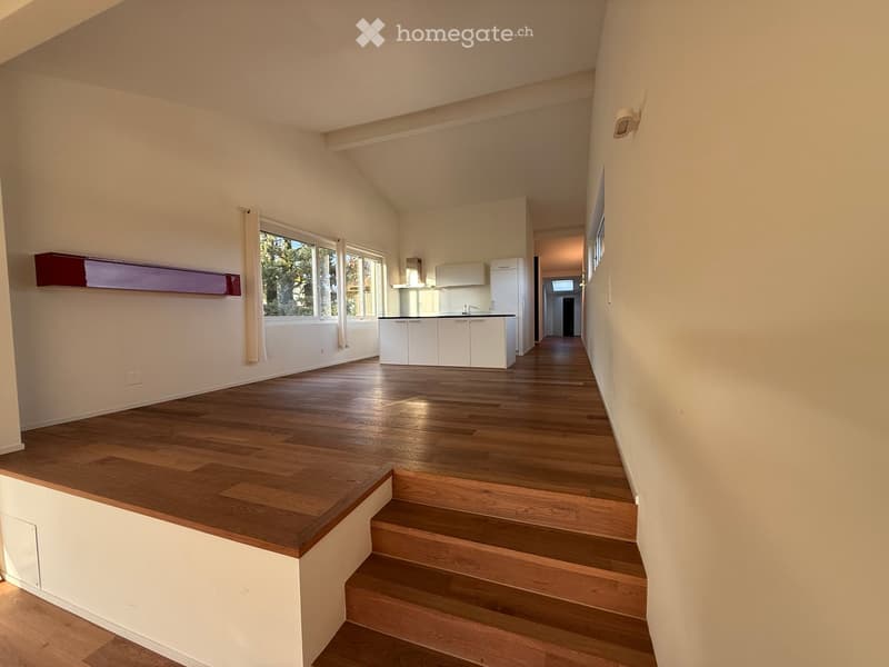 Bright, open hallway with wooden flooring, leading to a kitchen area with white cabinets and a staircase ascending to an upper level.