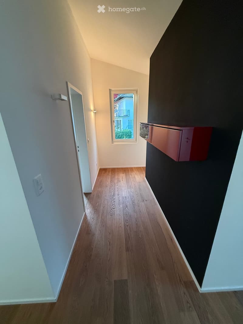 A narrow hallway with wooden flooring, white walls, and a window at the end showing a view of a building and greenery outside.