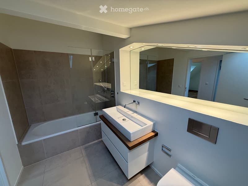 Modern bathroom with a white sink, wooden cabinet, large mirror, and bathtub with glass shower enclosure.