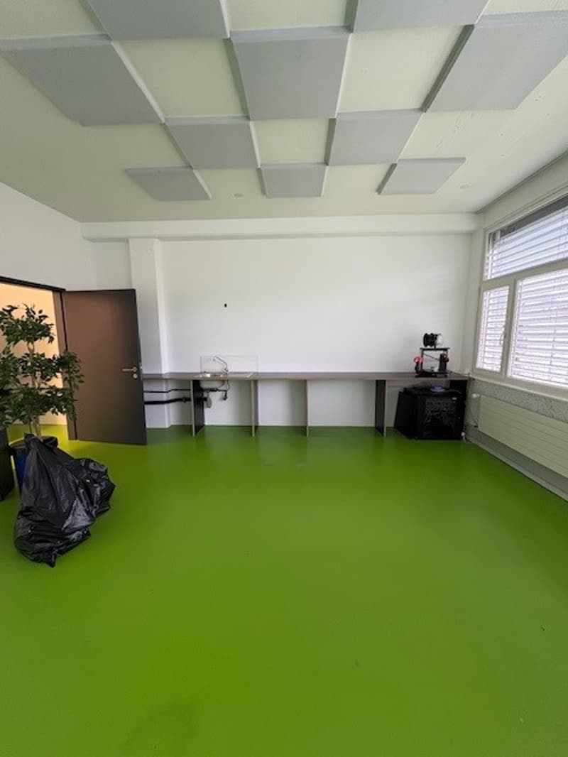 An empty room with white walls, green floor, ceiling panels, desk with faucet, potted plant, trash bag, windows with blinds, black object on desk