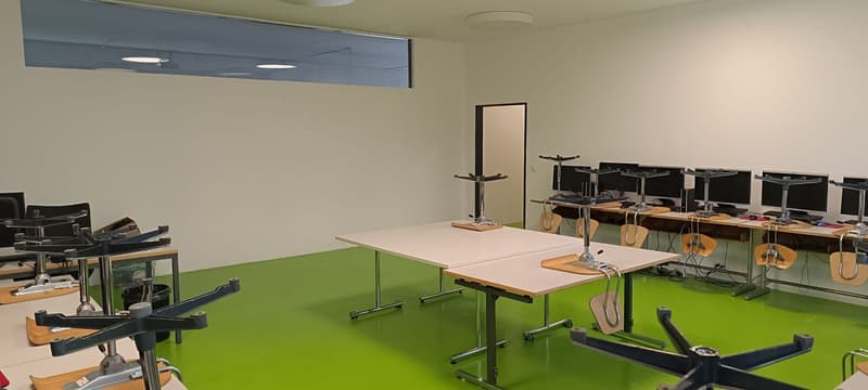 A room with white walls, green floor, multiple desks, monitors, and chairs.