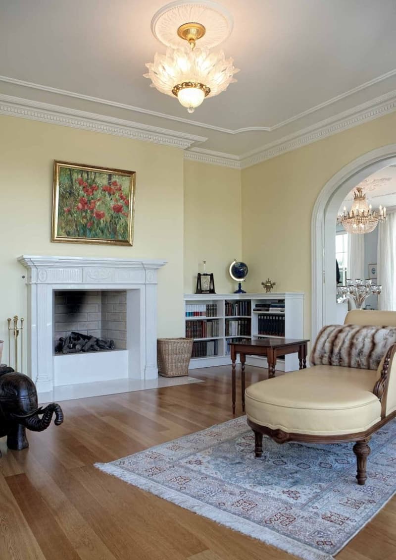 This image shows a spacious and elegant living room with a fireplace, hardwood floors, and a decorative chandelier. The room features a large area rug, a plush armchair, and built-in bookshelves. The walls are painted in a light, neutral color, and there