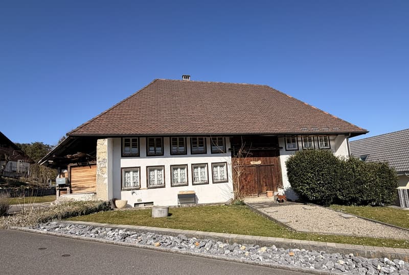 white house, brown roof, wooden door, multiple windows, gravel driveway, small garden