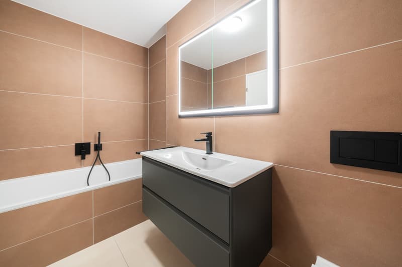 Modern bathroom with beige tile walls, a black vanity cabinet with a white sink, a large mirror with LED lighting, and a bathtub.