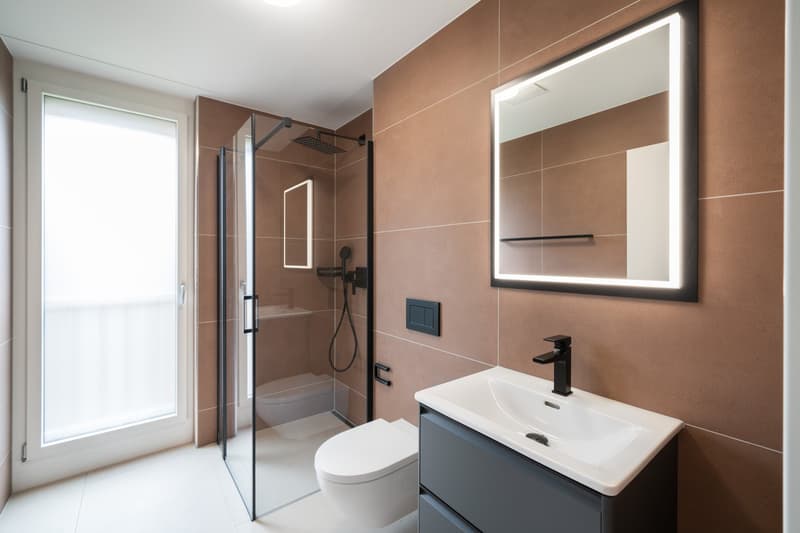 Modern bathroom with beige tile walls, large mirror with lighting, glass shower enclosure, vanity with sink, and toilet