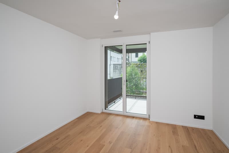 Spacious bedroom with hardwood flooring, white walls, and a sliding glass door leading to a balcony or terrace overlooking greenery outside.