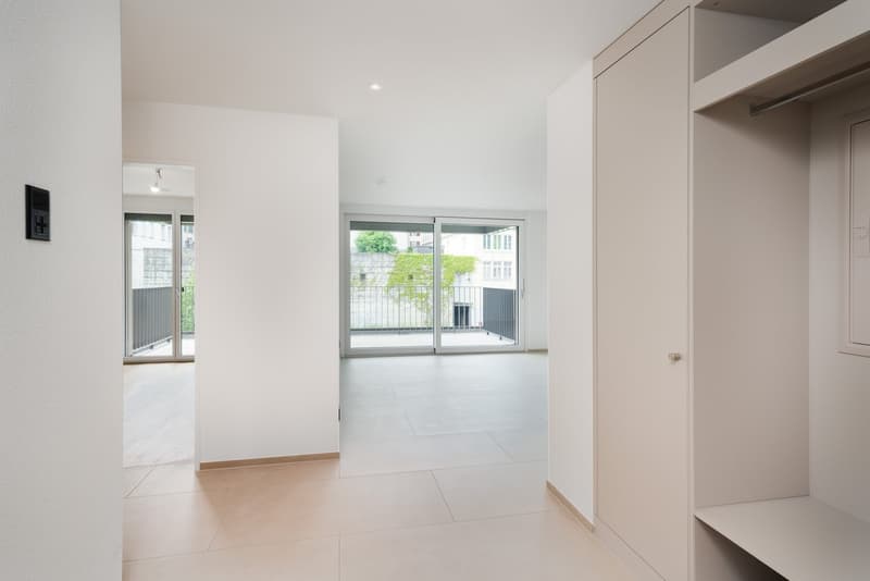 Spacious open-plan living area with large windows, sliding glass doors leading to a balcony, white walls, and light-colored tile flooring.