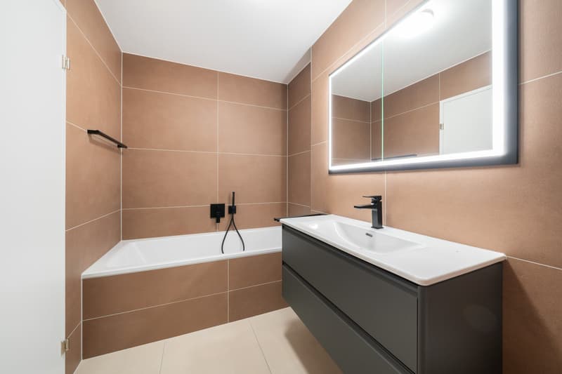 Modern bathroom with beige tile walls, a white bathtub, a vanity with a sink, and a large mirror with LED lighting