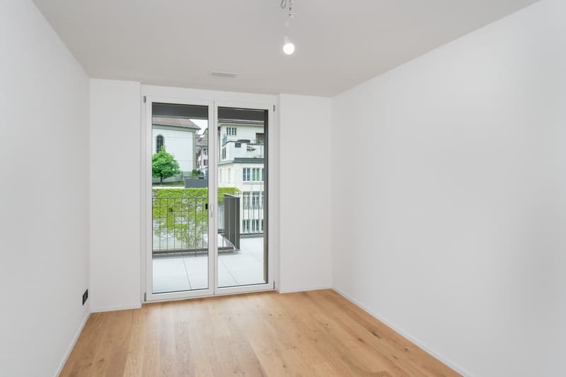 Spacious room with large windows, hardwood flooring, and access to a balcony or terrace overlooking the surrounding buildings and greenery.