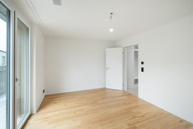 Spacious bedroom with large windows, hardwood flooring, and a white door leading to another room.