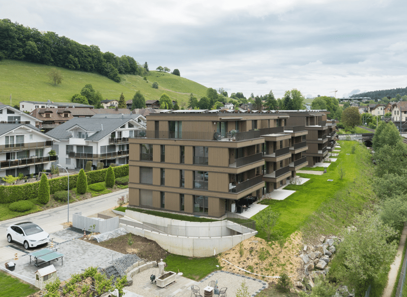 Multi-story apartment building with balconies, surrounded by green hills and trees, with a paved area and some vehicles visible in the foreground.